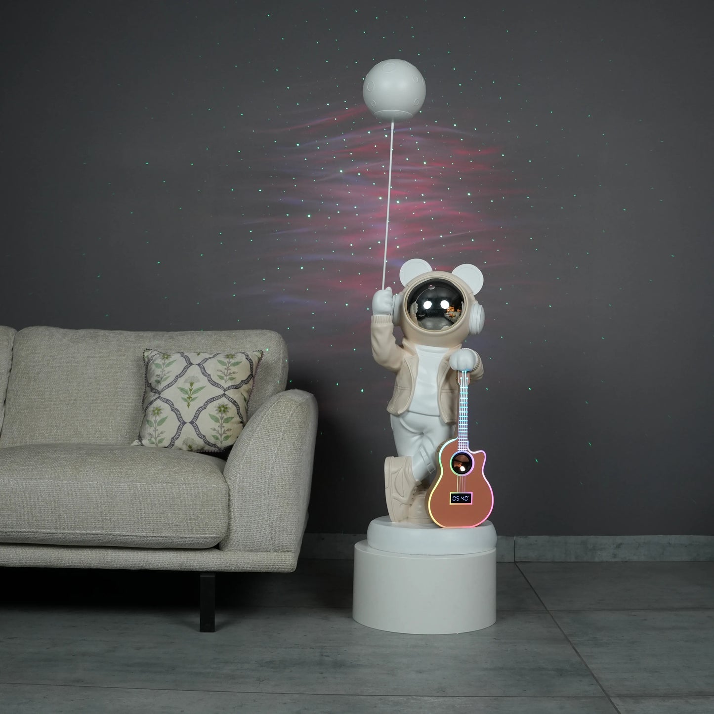 Beige and white astronaut statue with a chrome silver visor holding a moon-shaped projector balloon and a silver-tone guitar Bluetooth speaker, standing on a pedestal.