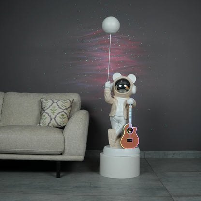 Beige and white astronaut statue with a chrome silver visor holding a moon-shaped projector balloon and a silver-tone guitar Bluetooth speaker, standing on a pedestal.