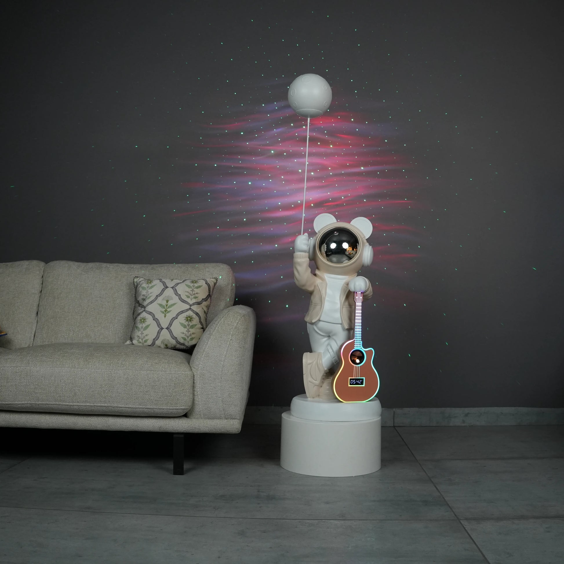 Beige and white astronaut statue with a chrome silver visor holding a moon-shaped projector balloon and a silver-tone guitar Bluetooth speaker, standing on a pedestal.