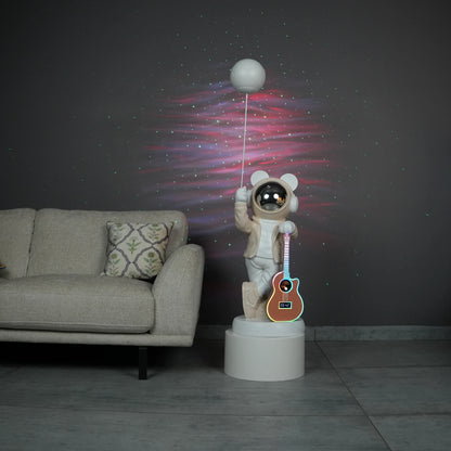 Beige and white astronaut statue with a chrome silver visor holding a moon-shaped projector balloon and a silver-tone guitar Bluetooth speaker, standing on a pedestal.