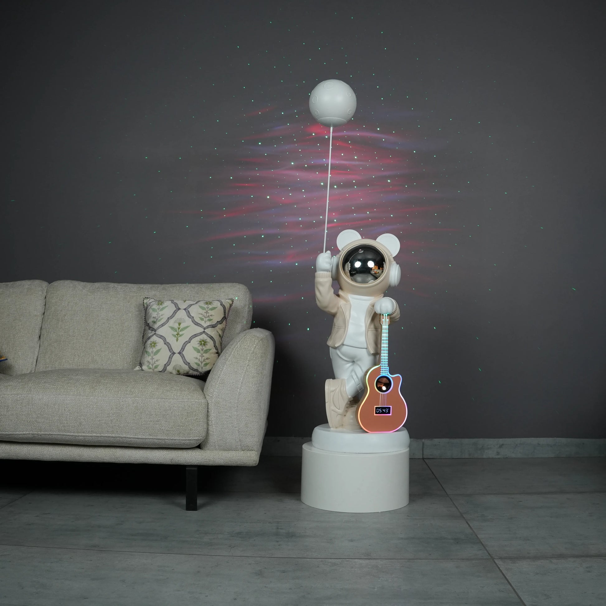 Beige and white astronaut statue with a chrome silver visor holding a moon-shaped projector balloon and a silver-tone guitar Bluetooth speaker, standing on a pedestal.