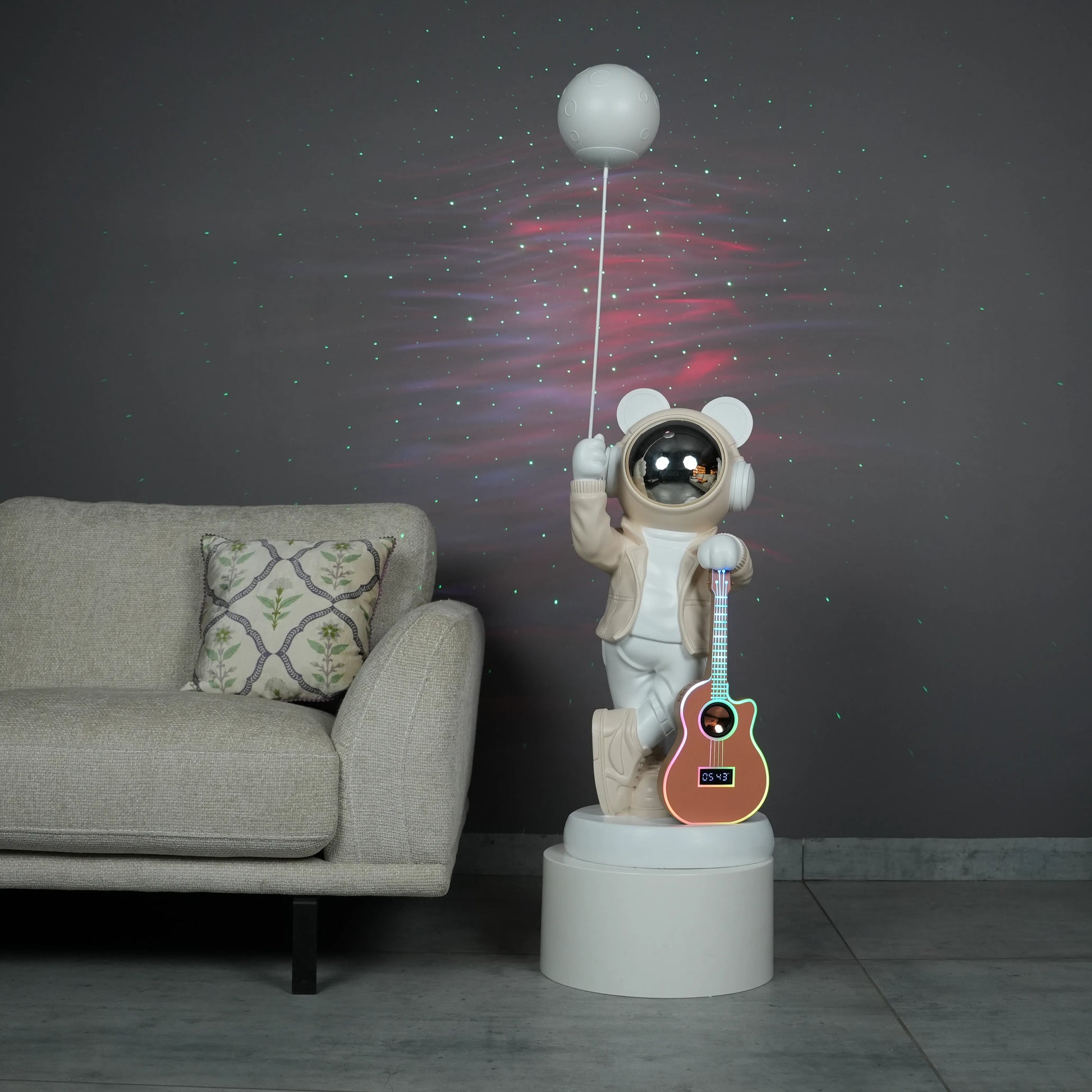 Beige and white astronaut statue with a chrome silver visor holding a moon-shaped projector balloon and a silver-tone guitar Bluetooth speaker, standing on a pedestal.