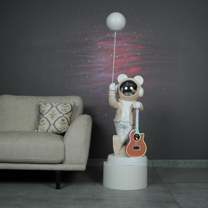 Beige and white astronaut statue with a chrome silver visor holding a moon-shaped projector balloon and a silver-tone guitar Bluetooth speaker, standing on a pedestal.