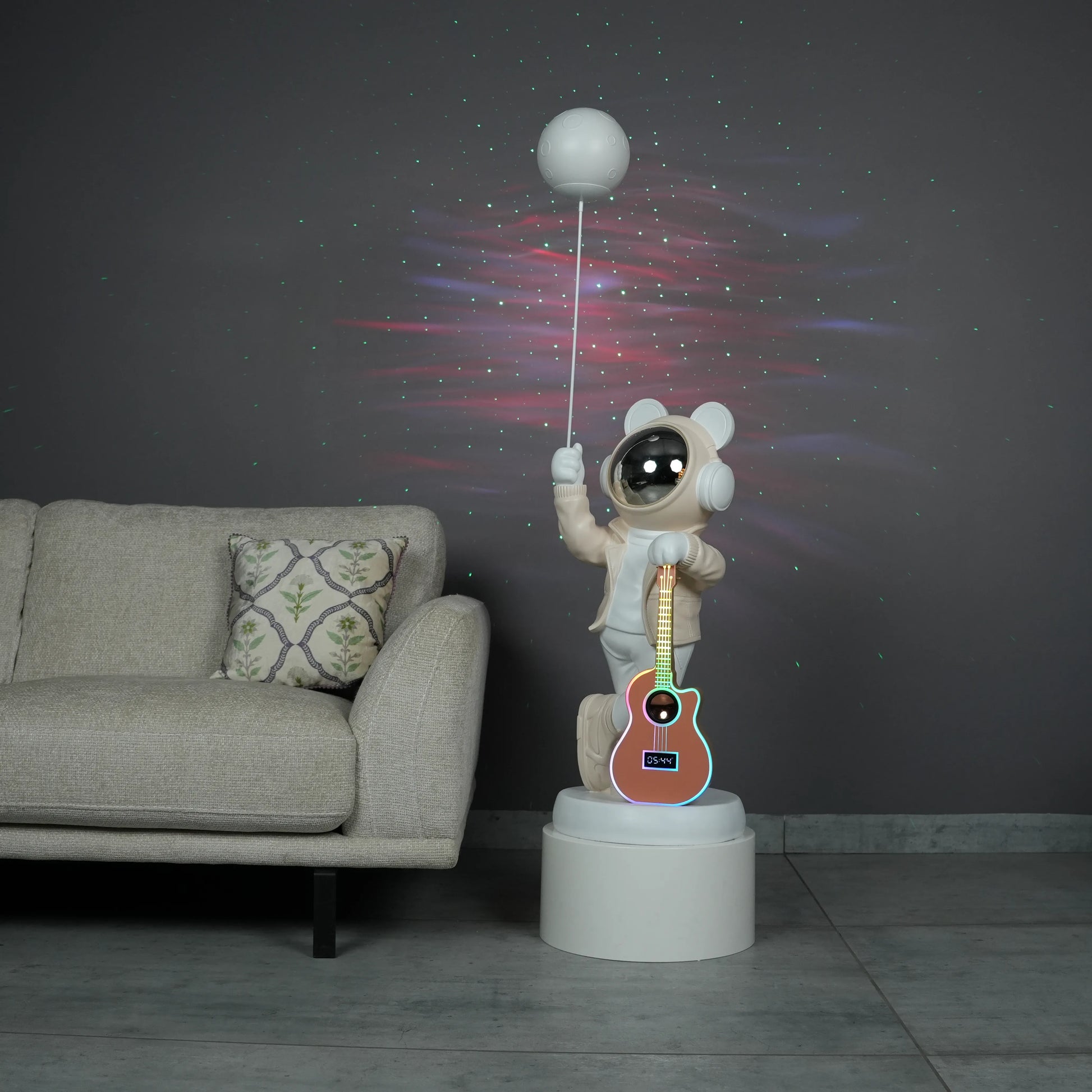 Beige and white astronaut statue with a chrome silver visor holding a moon-shaped projector balloon and a silver-tone guitar Bluetooth speaker, standing on a pedestal.