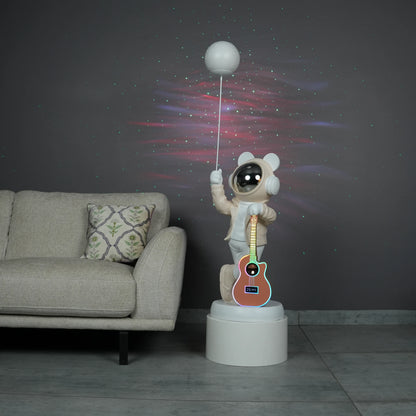 Beige and white astronaut statue with a chrome silver visor holding a moon-shaped projector balloon and a silver-tone guitar Bluetooth speaker, standing on a pedestal.