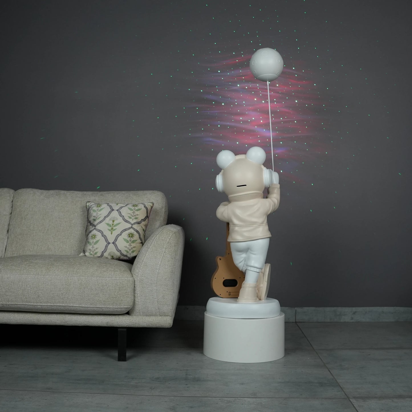 Beige and white astronaut statue with a chrome silver visor holding a moon-shaped projector balloon and a silver-tone guitar Bluetooth speaker, standing on a pedestal.