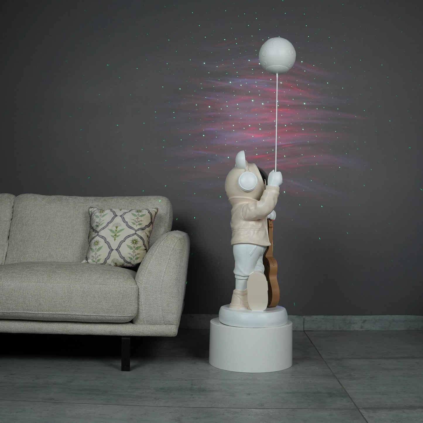 Beige and white astronaut statue with a chrome silver visor holding a moon-shaped projector balloon and a silver-tone guitar Bluetooth speaker, standing on a pedestal.