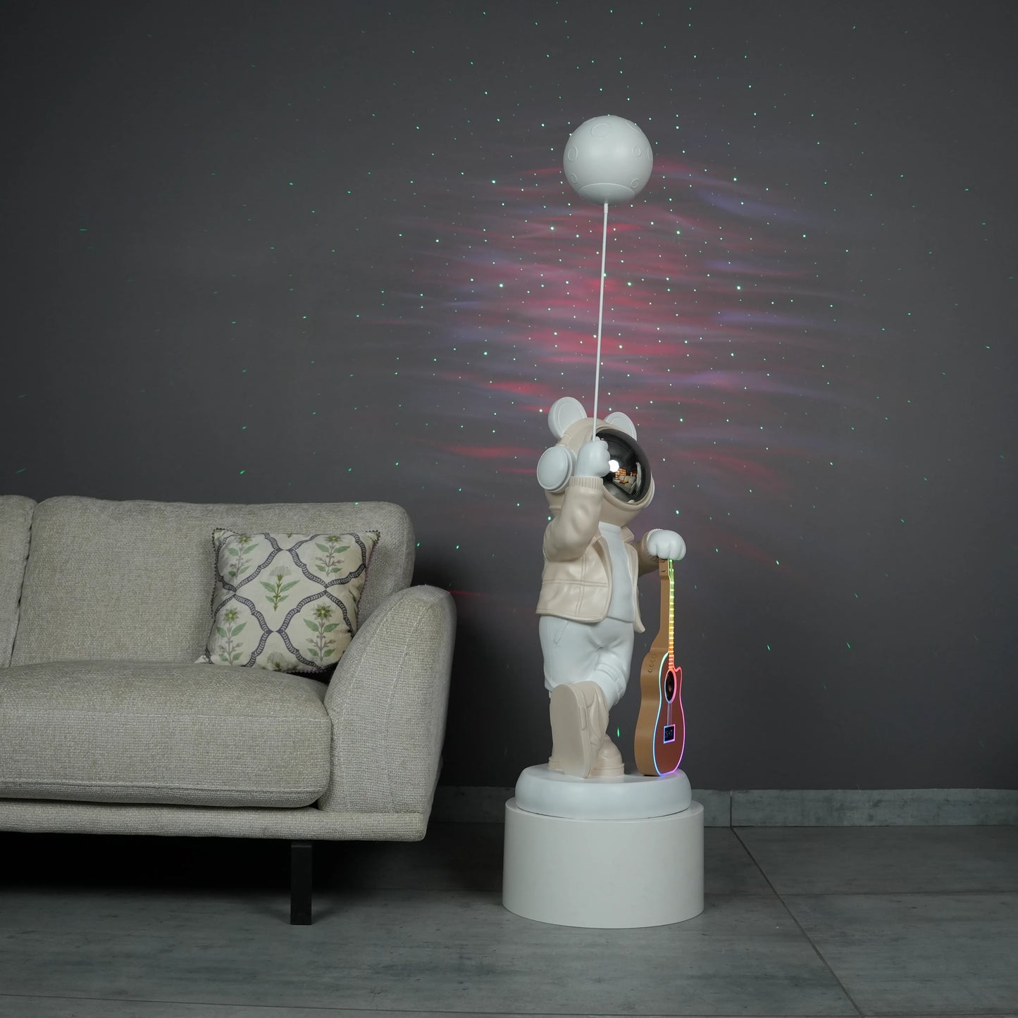 Beige and white astronaut statue with a chrome silver visor holding a moon-shaped projector balloon and a silver-tone guitar Bluetooth speaker, standing on a pedestal.