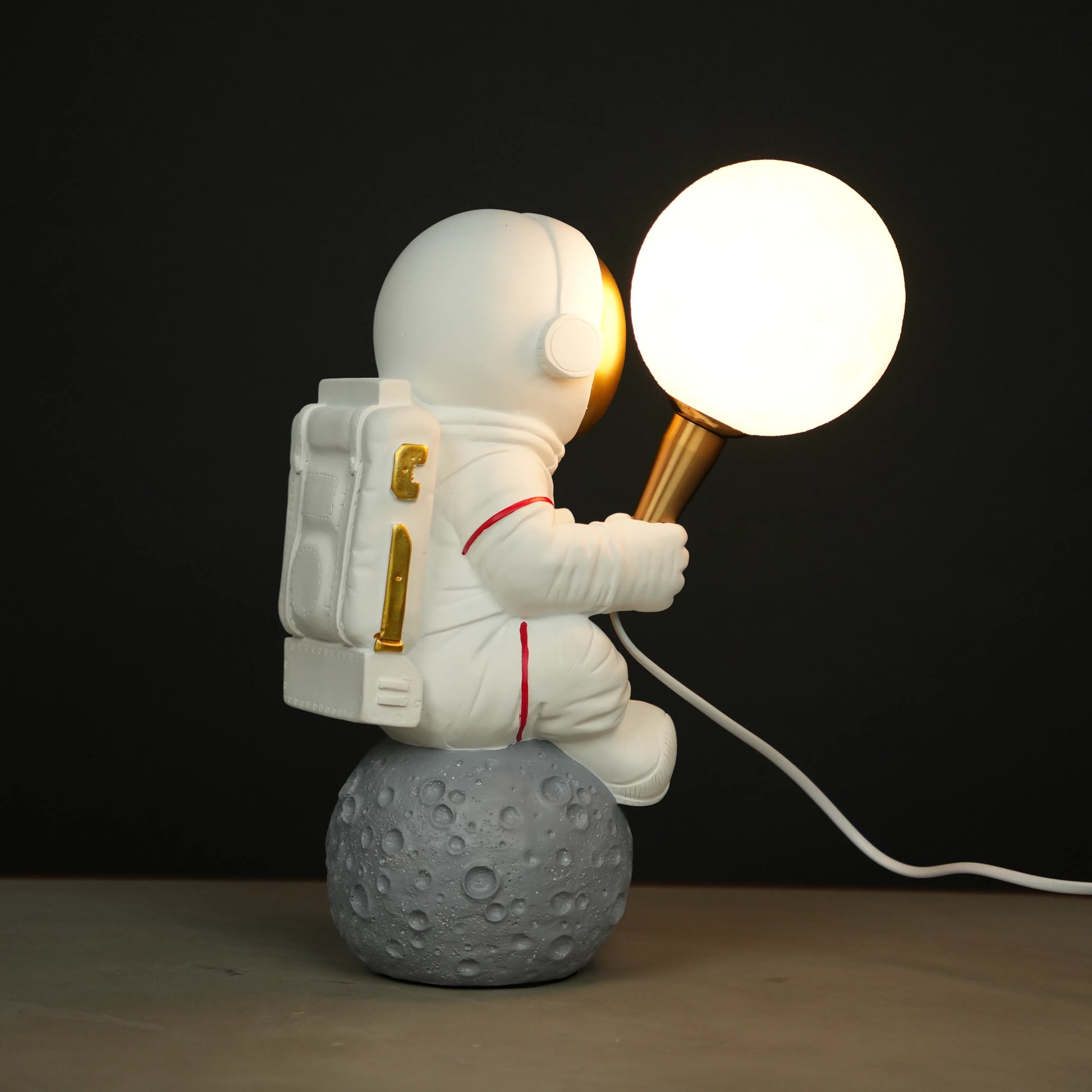 White astronaut figurine with a gold visor sitting on a textured grey moon while holding a glowing orb lamp, placed against a dark background.