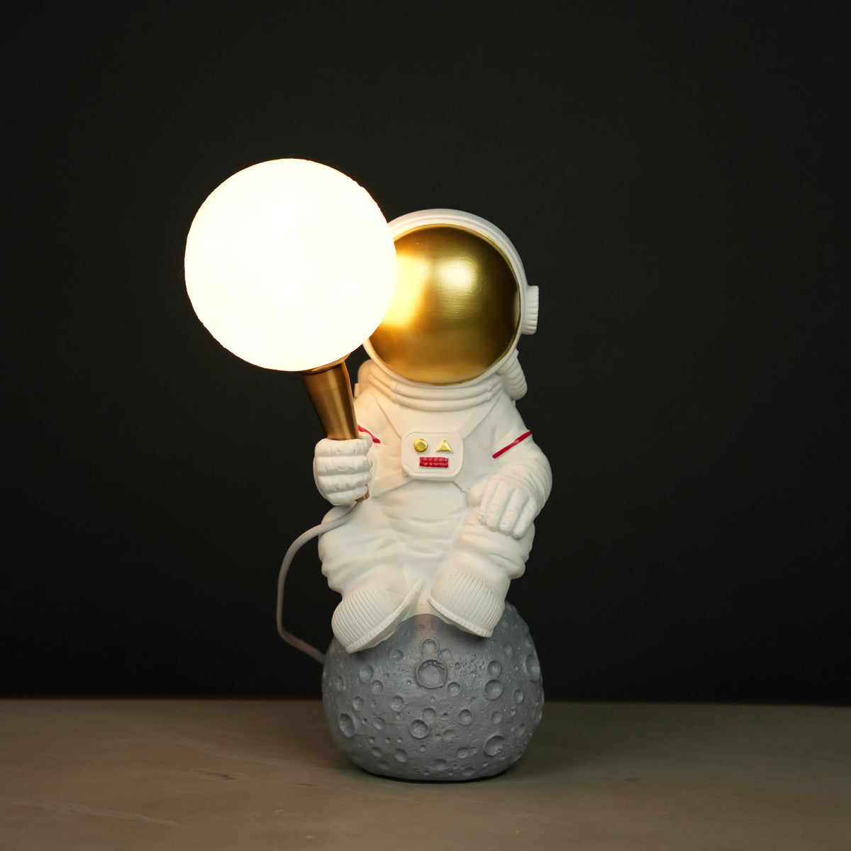 White astronaut figurine with a gold visor sitting on a textured grey moon while holding a glowing orb lamp, placed against a dark background.