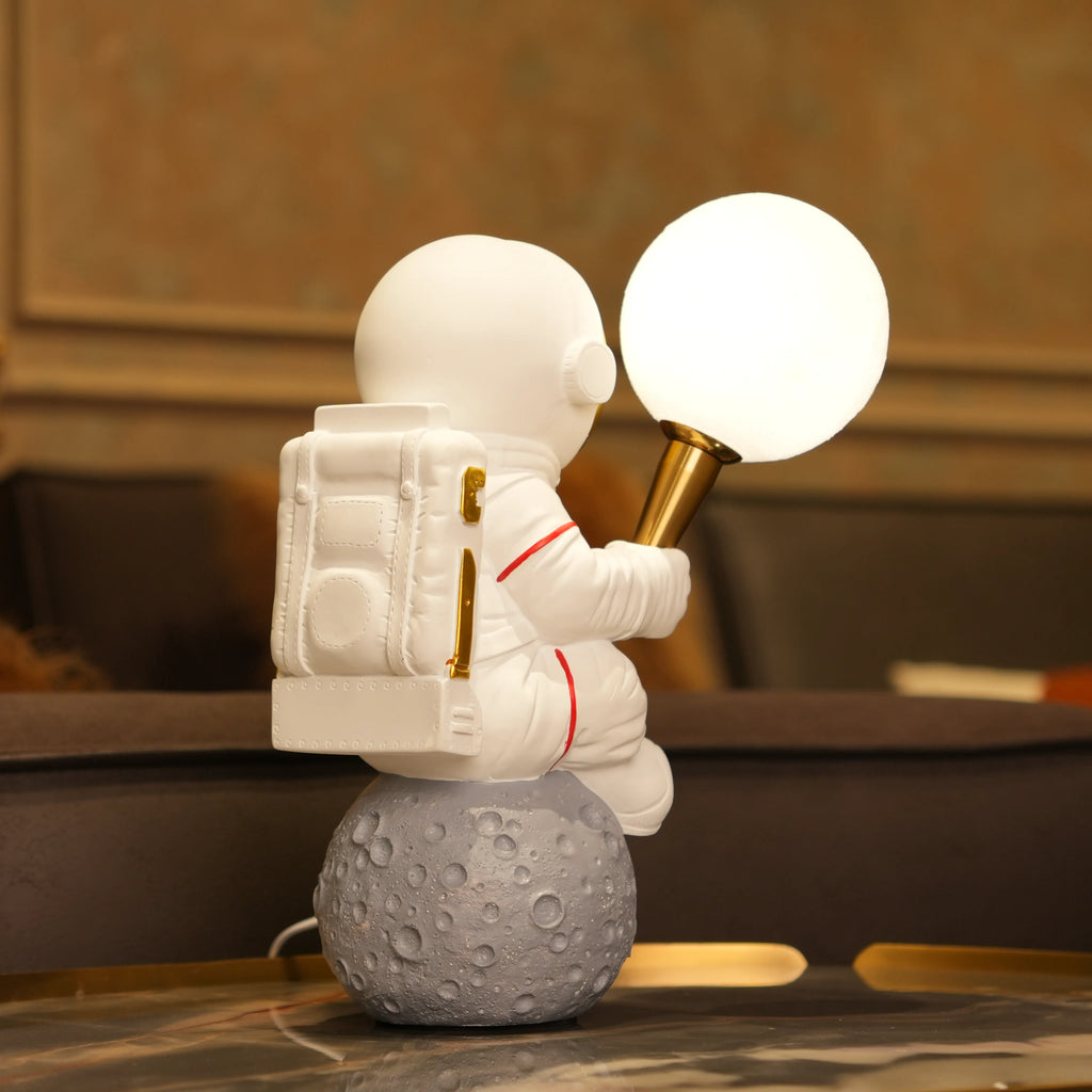 White astronaut figurine with a gold visor sitting on a textured grey moon while holding a glowing orb lamp, placed against a dark background.