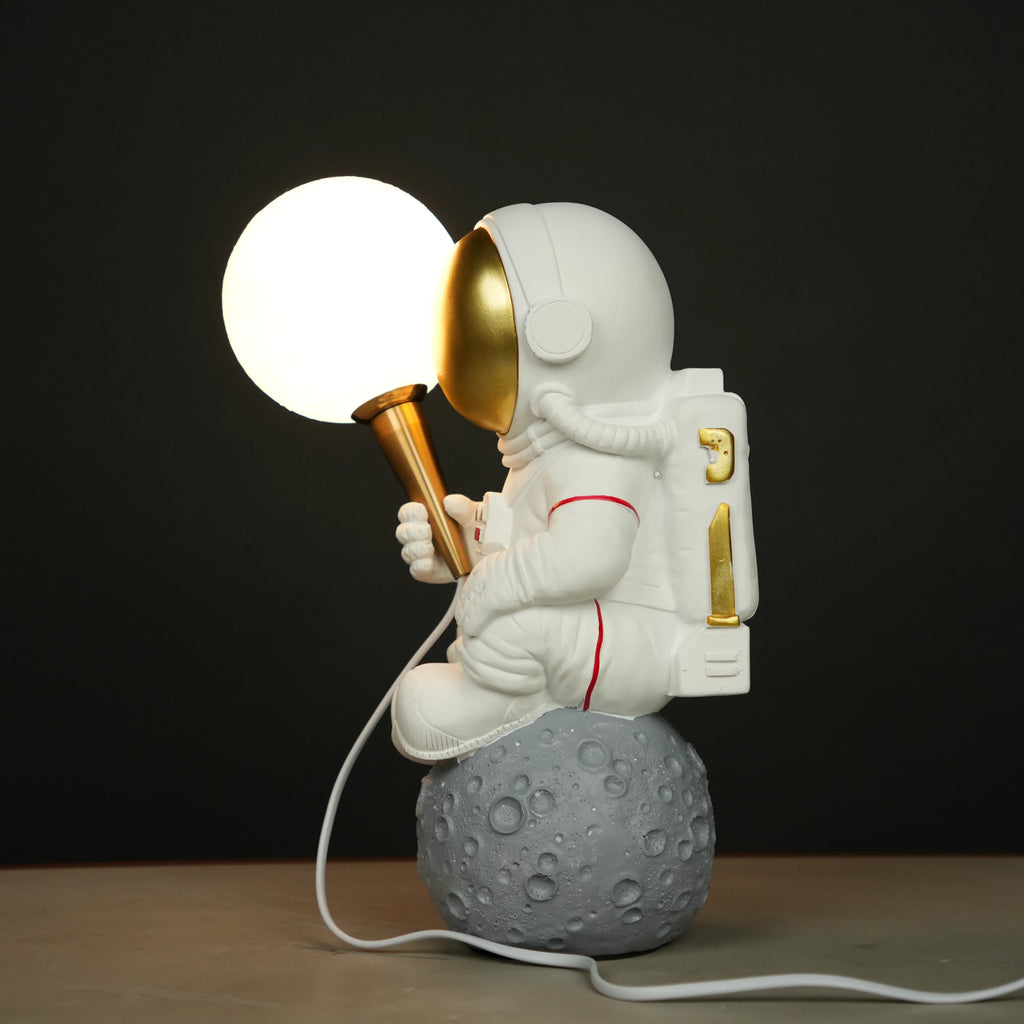 White astronaut figurine with a gold visor sitting on a textured grey moon while holding a glowing orb lamp, placed against a dark background.