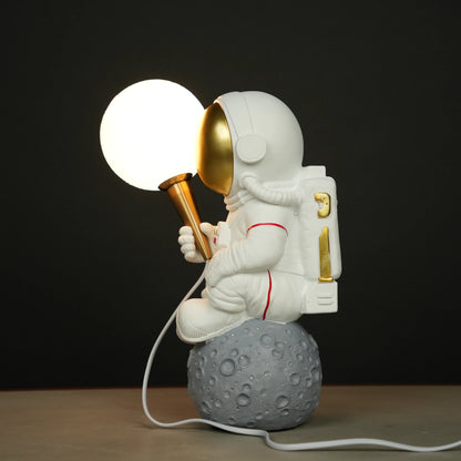 White astronaut figurine with a gold visor sitting on a textured grey moon while holding a glowing orb lamp, placed against a dark background.