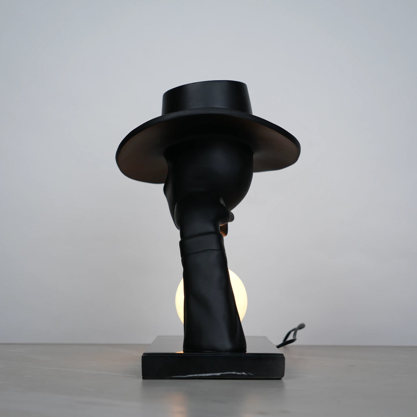 Elegant Hat Lamp – Matte black faceless figurine with wide hat holding glowing orb on marble base – Ghar Art.