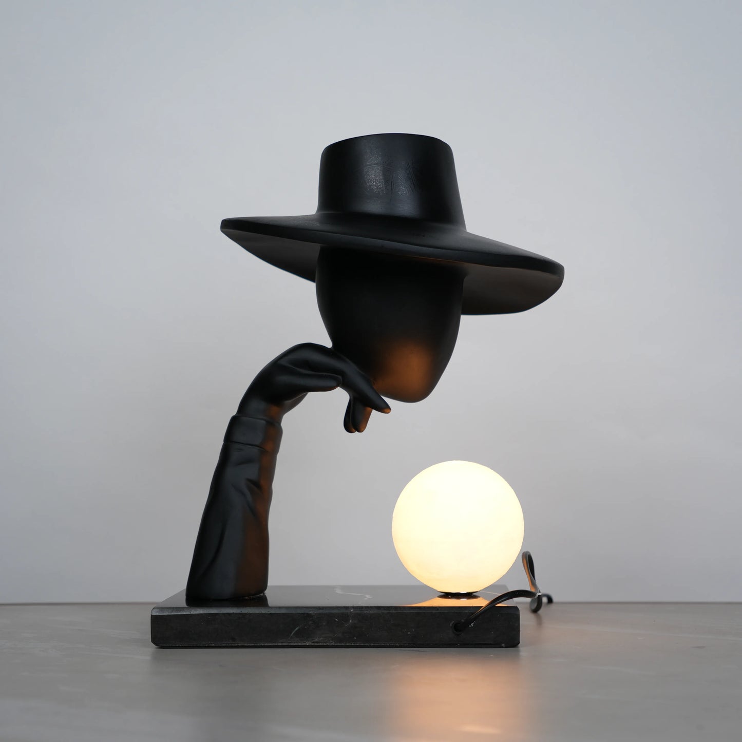 Elegant Hat Lamp – Matte black faceless figurine with wide hat holding glowing orb on marble base – Ghar Art.