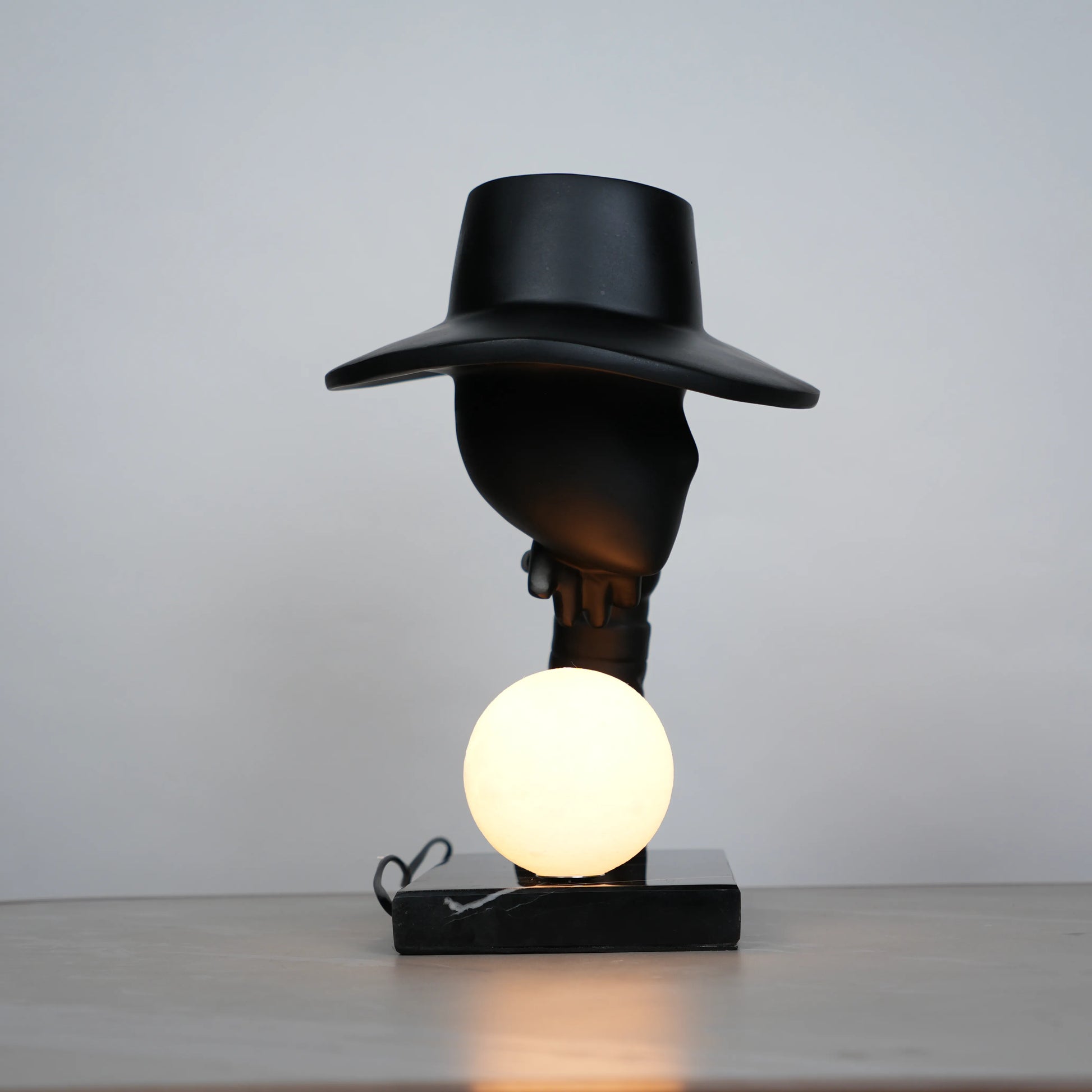 Elegant Hat Lamp – Matte black faceless figurine with wide hat holding glowing orb on marble base – Ghar Art.