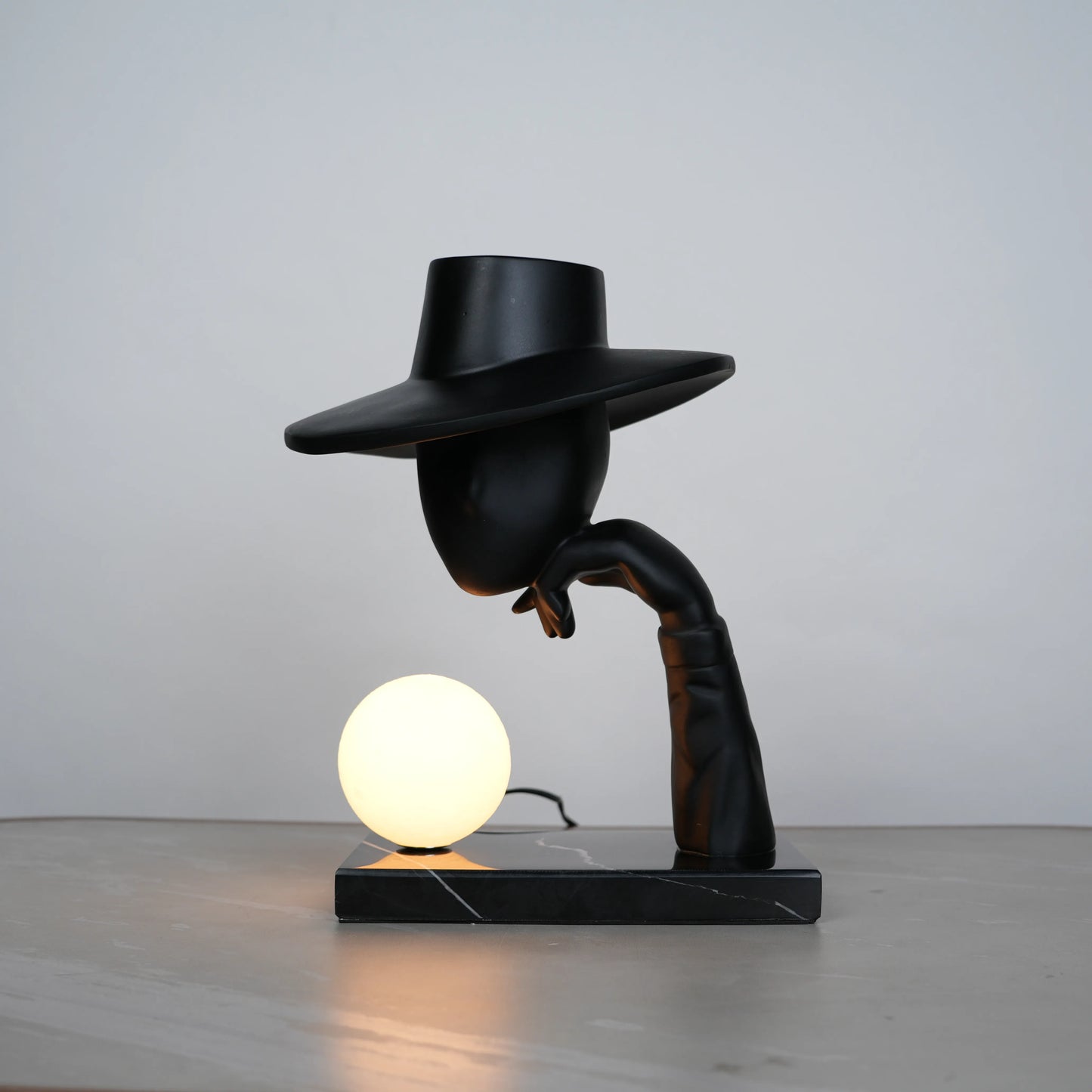 Elegant Hat Lamp – Matte black faceless figurine with wide hat holding glowing orb on marble base – Ghar Art.