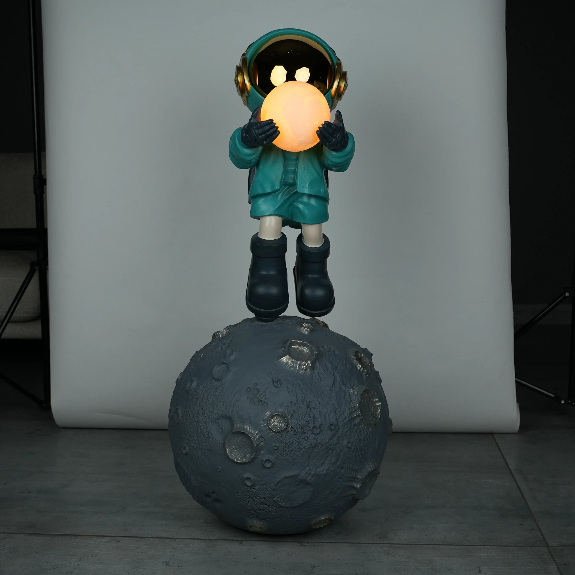 Teal astronaut figurine with gold visor holding a glowing orb while standing on a large crater-textured moon base.