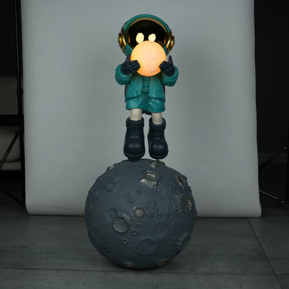 Teal astronaut figurine with gold visor holding a glowing orb while standing on a large crater-textured moon base.