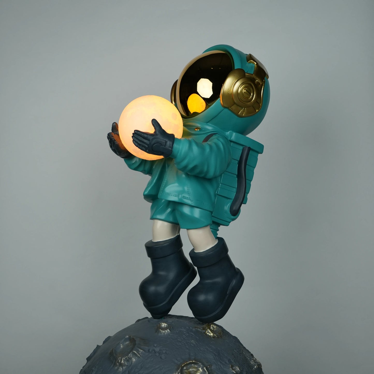 Teal astronaut figurine with gold visor holding a glowing orb while standing on a large crater-textured moon base.