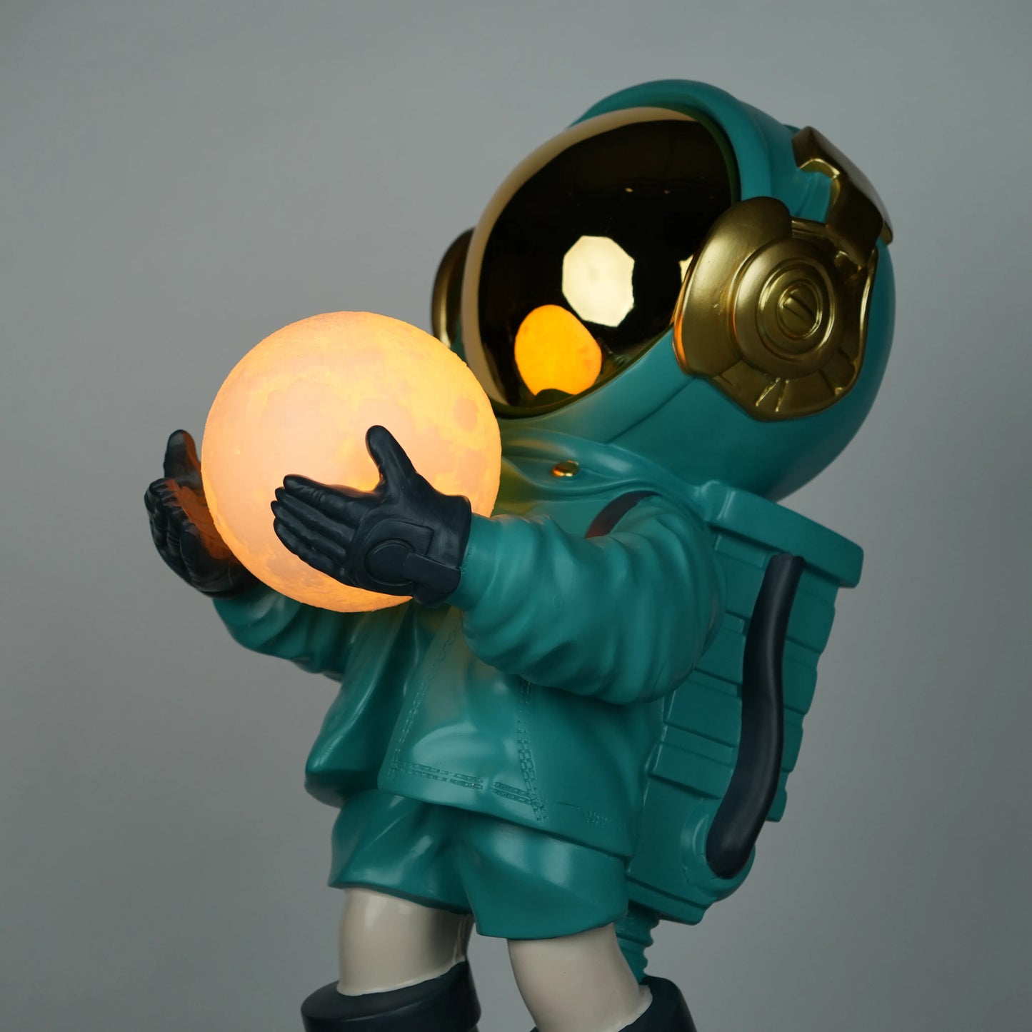Teal astronaut figurine with gold visor holding a glowing orb while standing on a large crater-textured moon base.