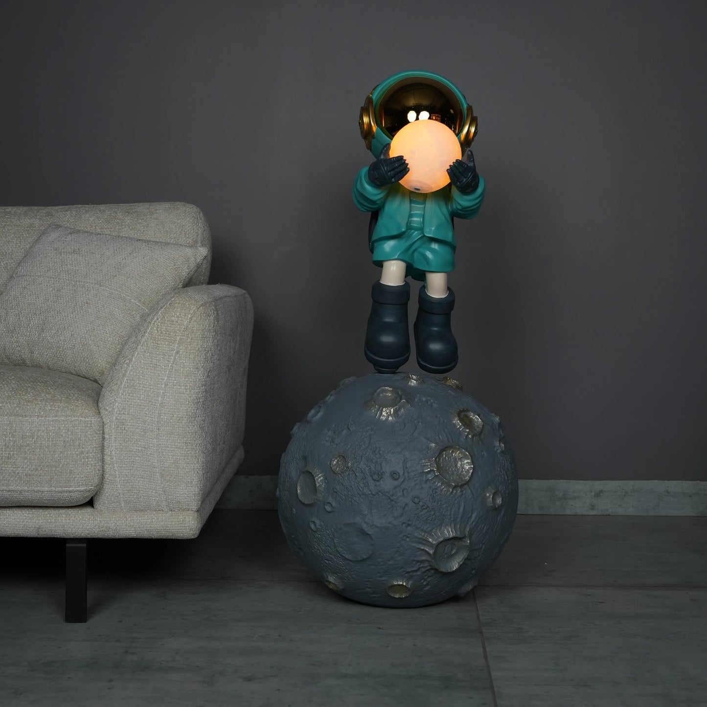 Teal astronaut figurine with gold visor holding a glowing orb while standing on a large crater-textured moon base.