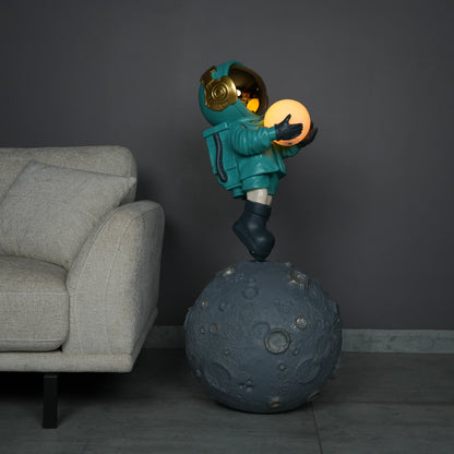 Teal astronaut figurine with gold visor holding a glowing orb while standing on a large crater-textured moon base.
