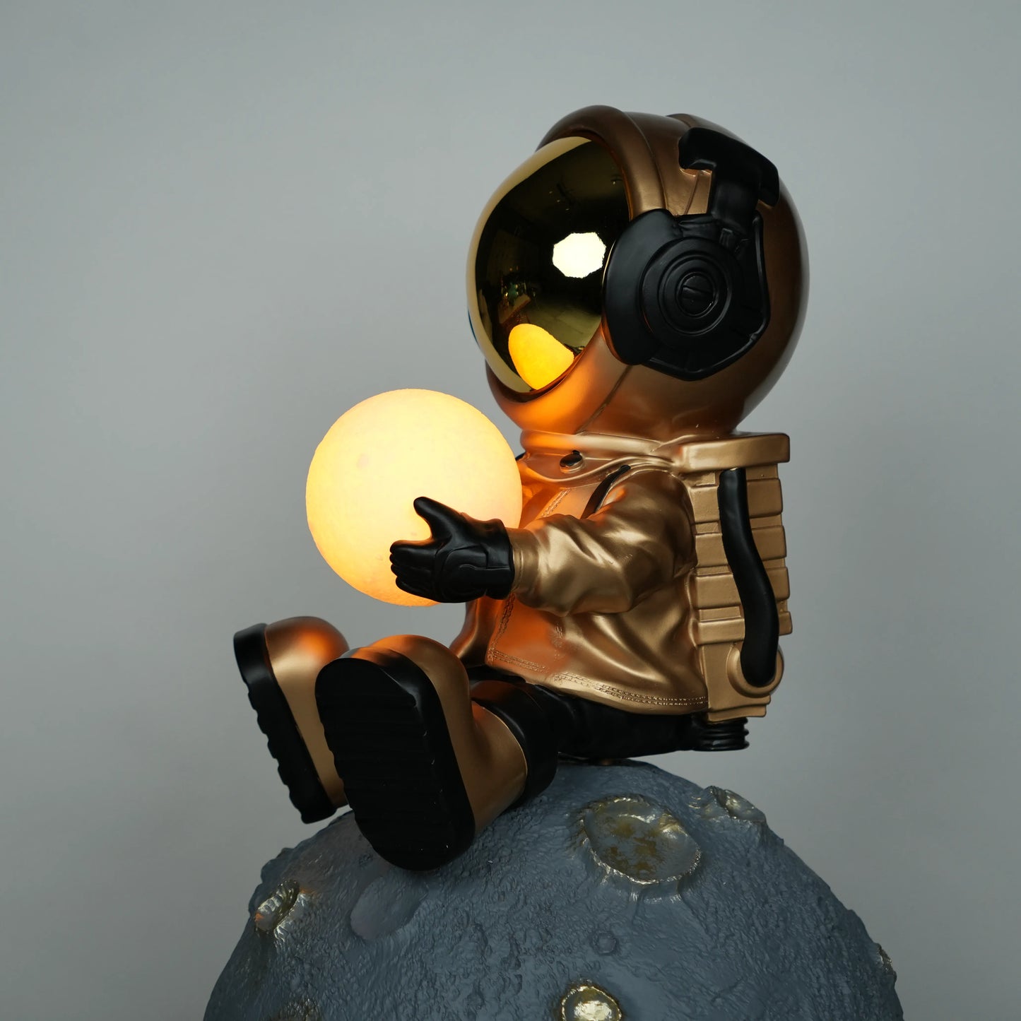 Gold astronaut figurine sitting on a large cratered moon, holding a glowing orb lamp with a chrome-gold visor.