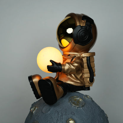 Gold astronaut figurine sitting on a large cratered moon, holding a glowing orb lamp with a chrome-gold visor.