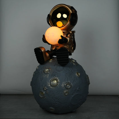 Gold astronaut figurine sitting on a large cratered moon, holding a glowing orb lamp with a chrome-gold visor.