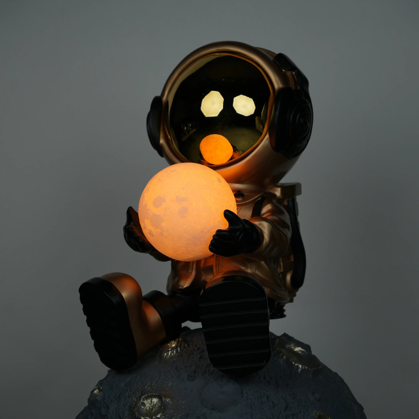 Gold astronaut figurine sitting on a large cratered moon, holding a glowing orb lamp with a chrome-gold visor.