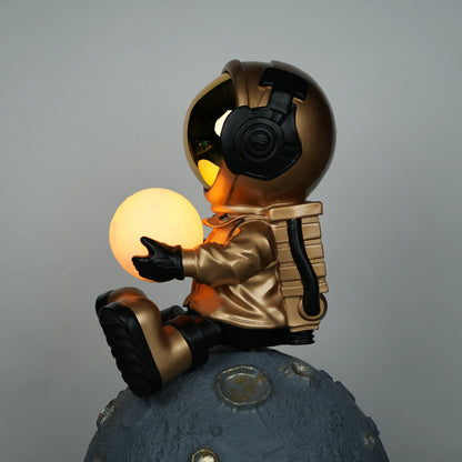 Gold astronaut figurine sitting on a large cratered moon, holding a glowing orb lamp with a chrome-gold visor.