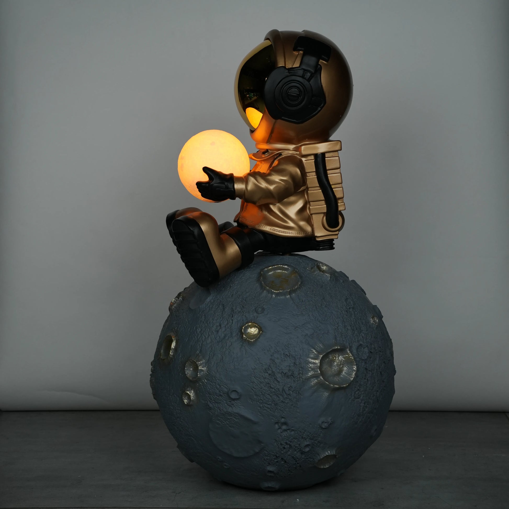 Gold astronaut figurine sitting on a large cratered moon, holding a glowing orb lamp with a chrome-gold visor.