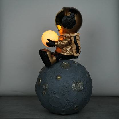 Gold astronaut figurine sitting on a large cratered moon, holding a glowing orb lamp with a chrome-gold visor.