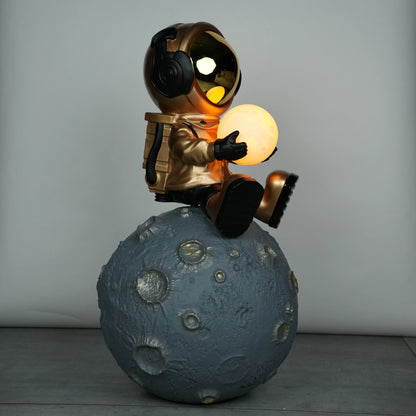 Gold astronaut figurine sitting on a large cratered moon, holding a glowing orb lamp with a chrome-gold visor.