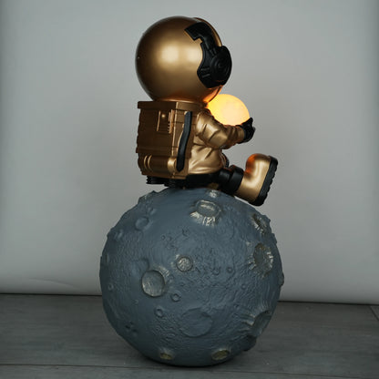 Gold astronaut figurine sitting on a large cratered moon, holding a glowing orb lamp with a chrome-gold visor.
