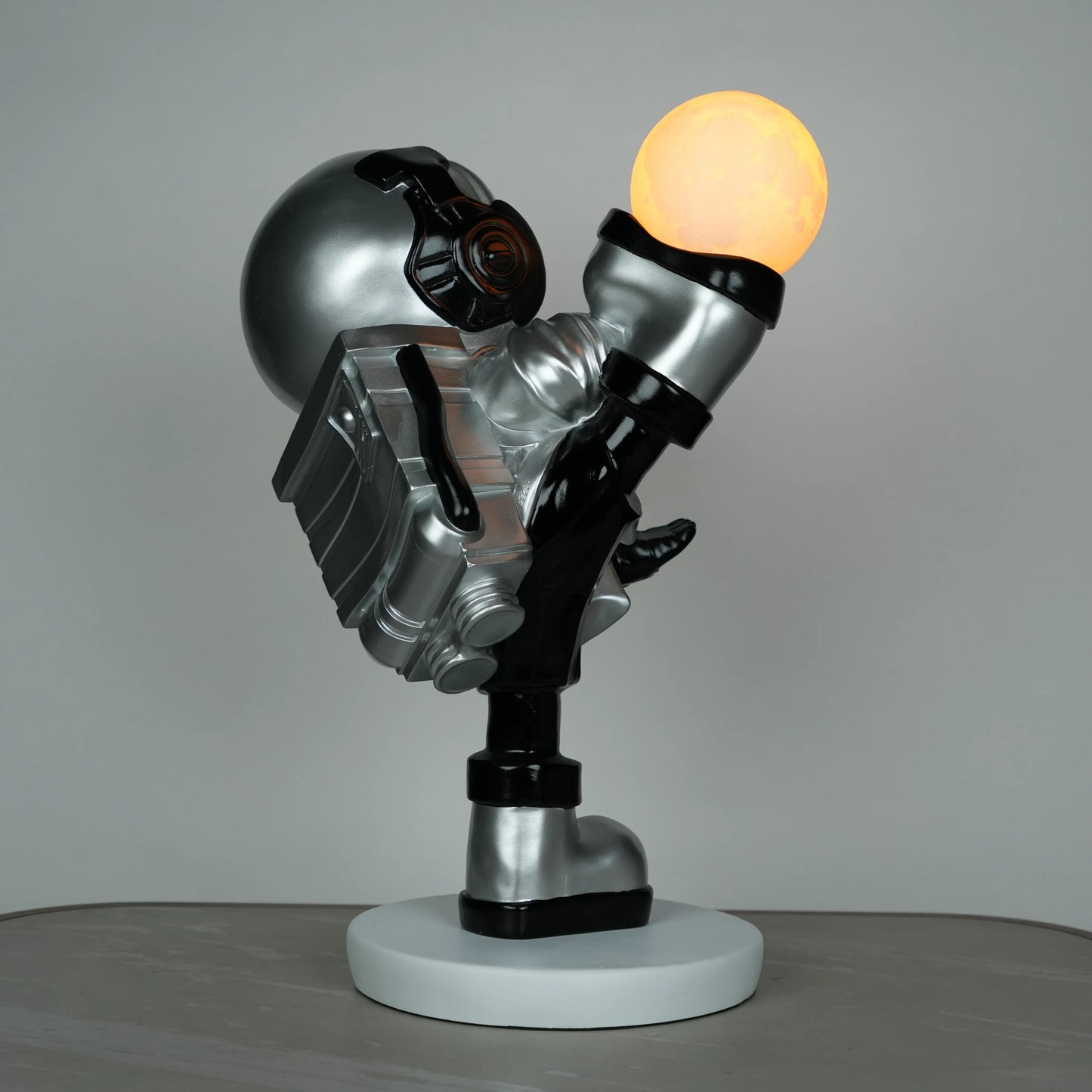 Silver astronaut lamp in a high-kick pose with chrome-gold visor and glowing ambient orb on white base; luxury modern décor lamp.