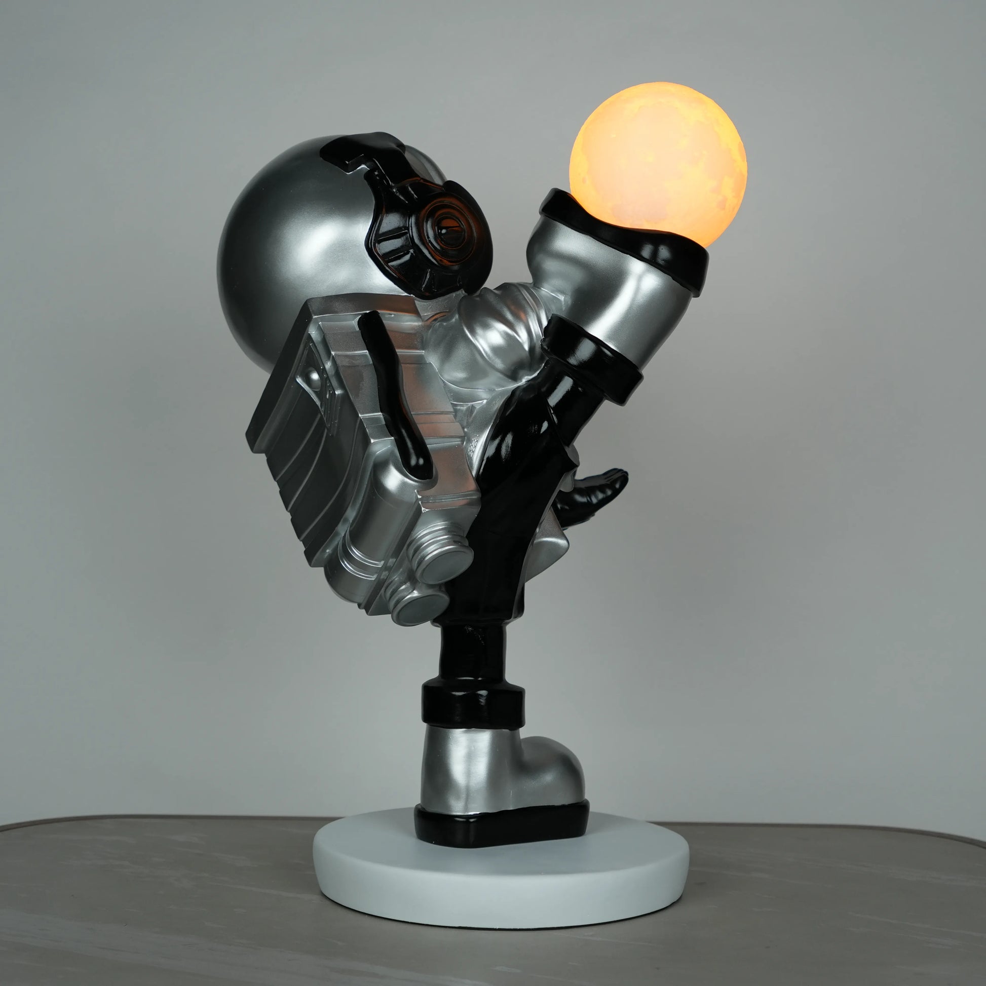 Silver astronaut lamp in a high-kick pose with chrome-gold visor and glowing ambient orb on white base; luxury modern décor lamp.