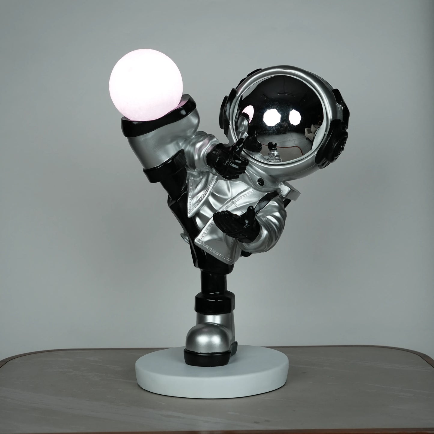 Silver astronaut lamp in a high-kick pose with chrome-gold visor and glowing ambient orb on white base; luxury modern décor lamp.