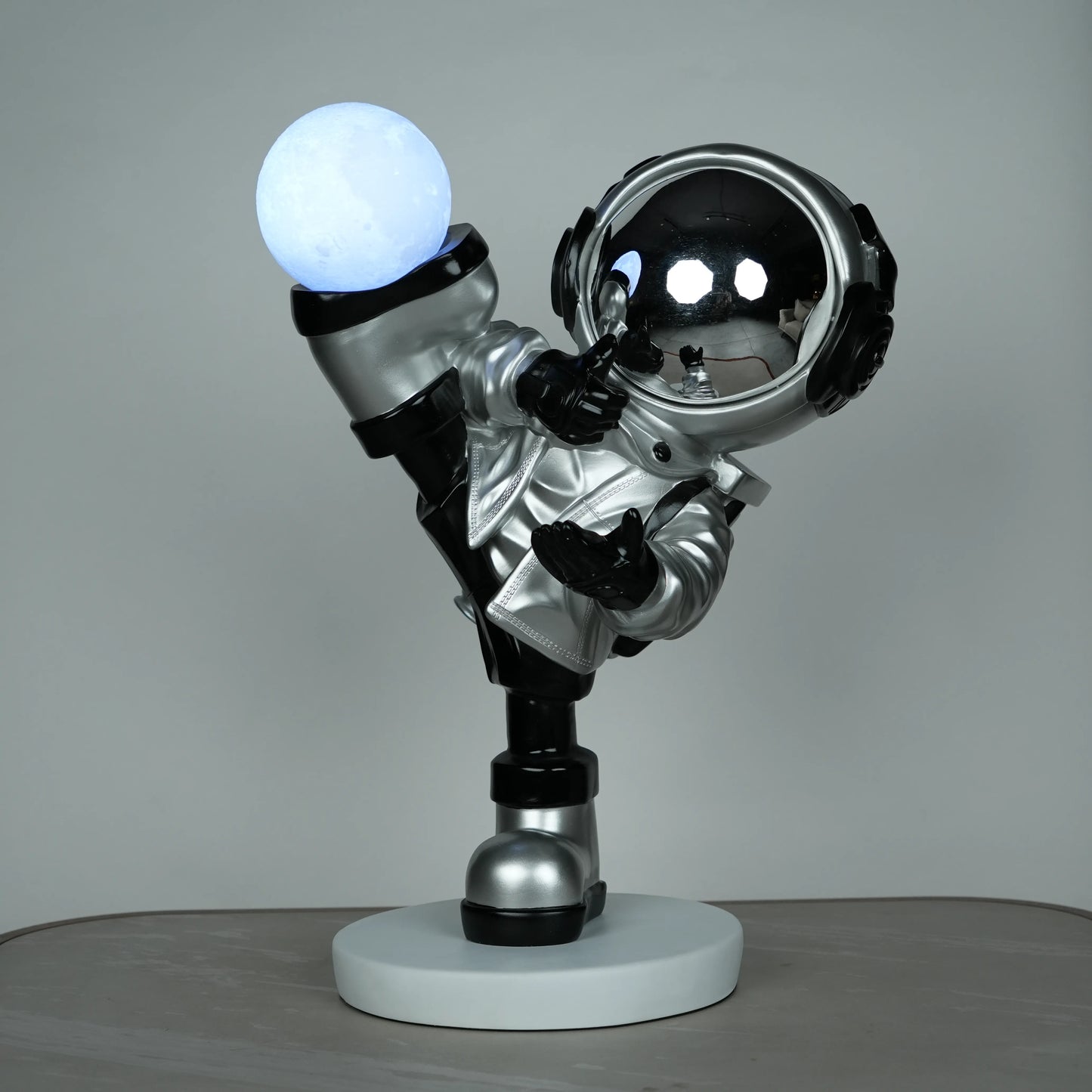 Silver astronaut lamp in a high-kick pose with chrome-gold visor and glowing ambient orb on white base; luxury modern décor lamp.