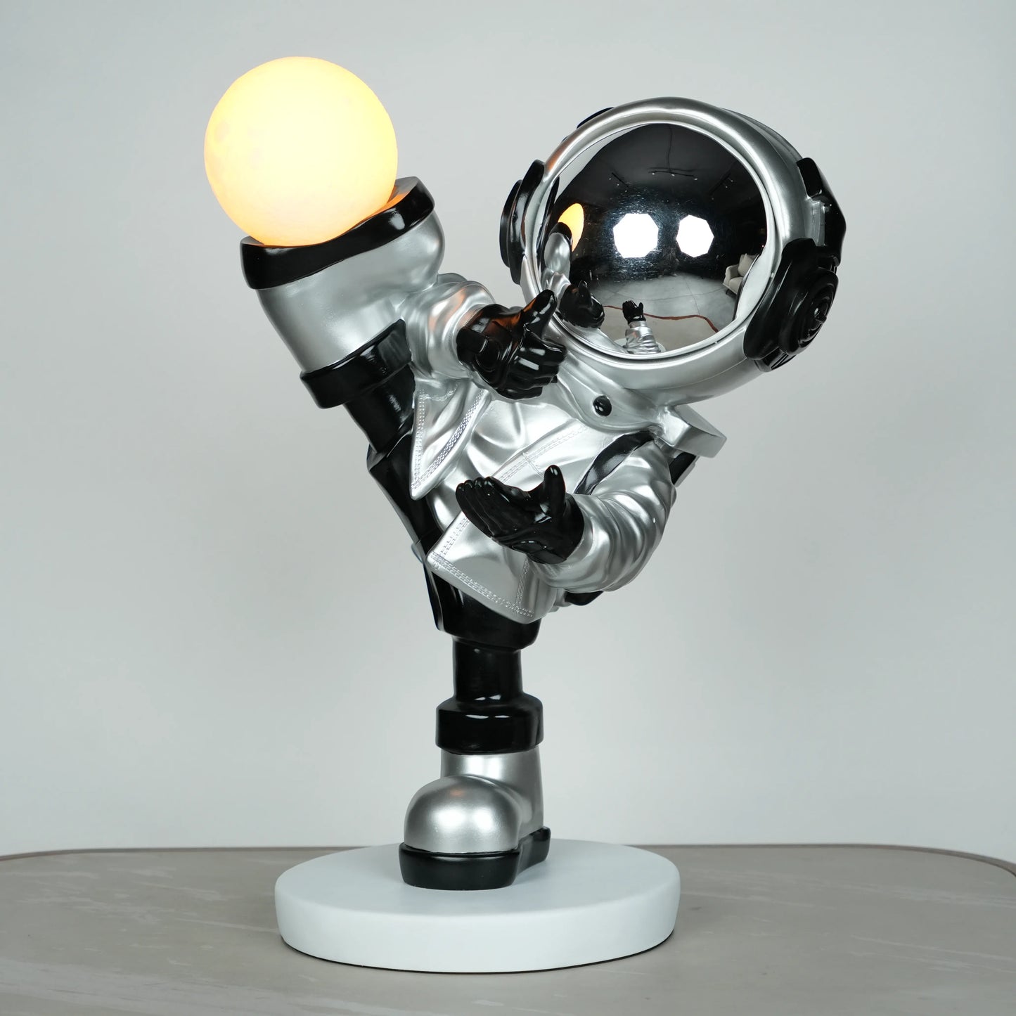 Silver astronaut lamp in a high-kick pose with chrome-gold visor and glowing ambient orb on white base; luxury modern décor lamp.