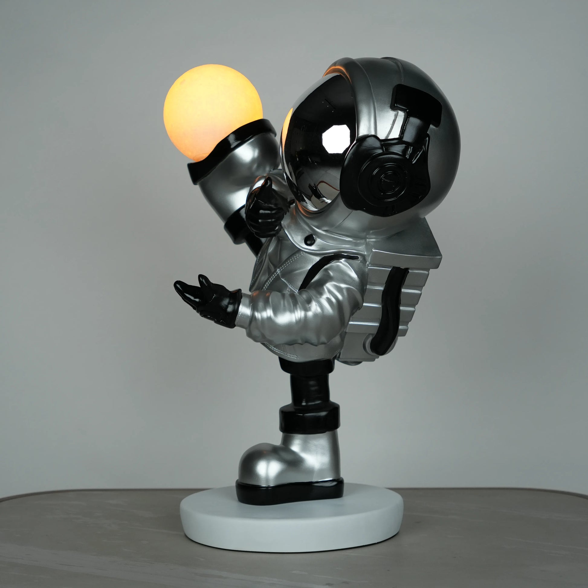 Silver astronaut lamp in a high-kick pose with chrome-gold visor and glowing ambient orb on white base; luxury modern décor lamp.