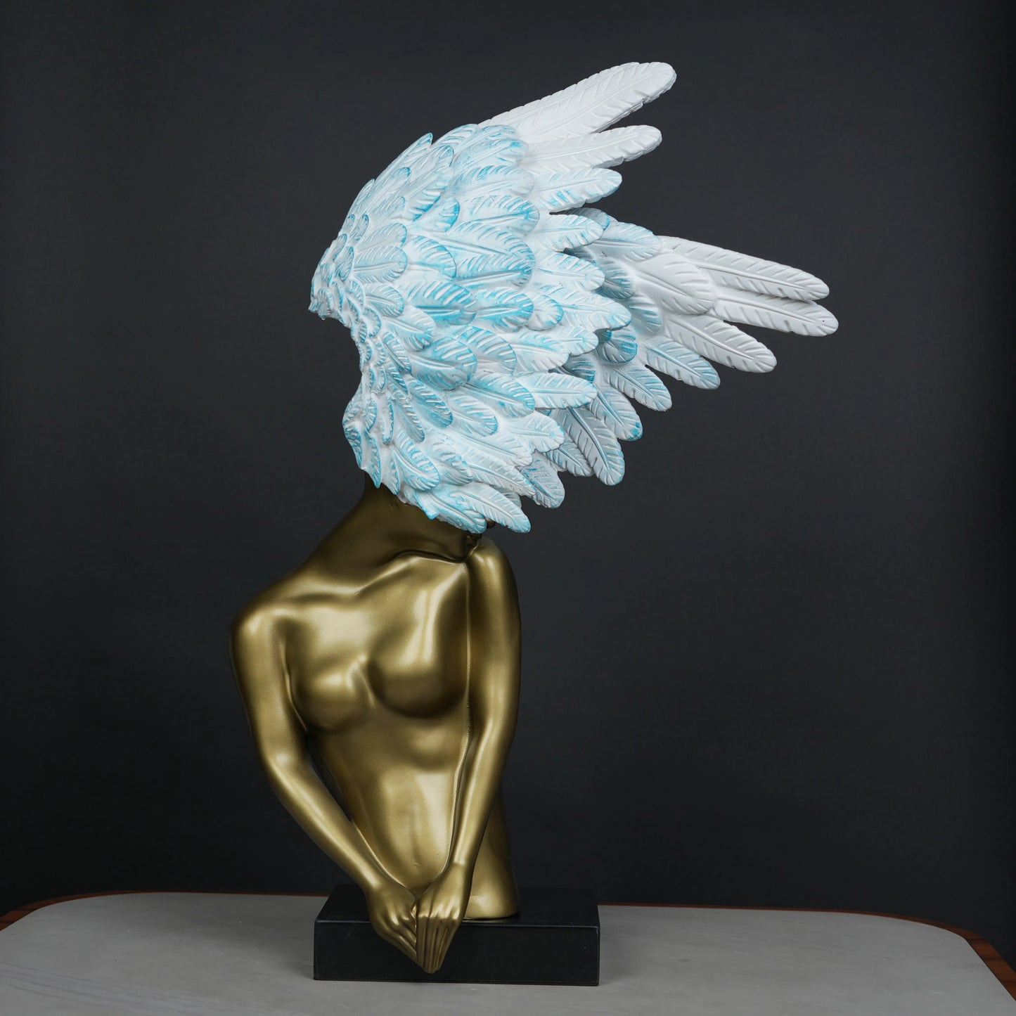 Gold-bodied woman sculpture with large white and blue feathered wings on a black base — luxury artistic décor piece.