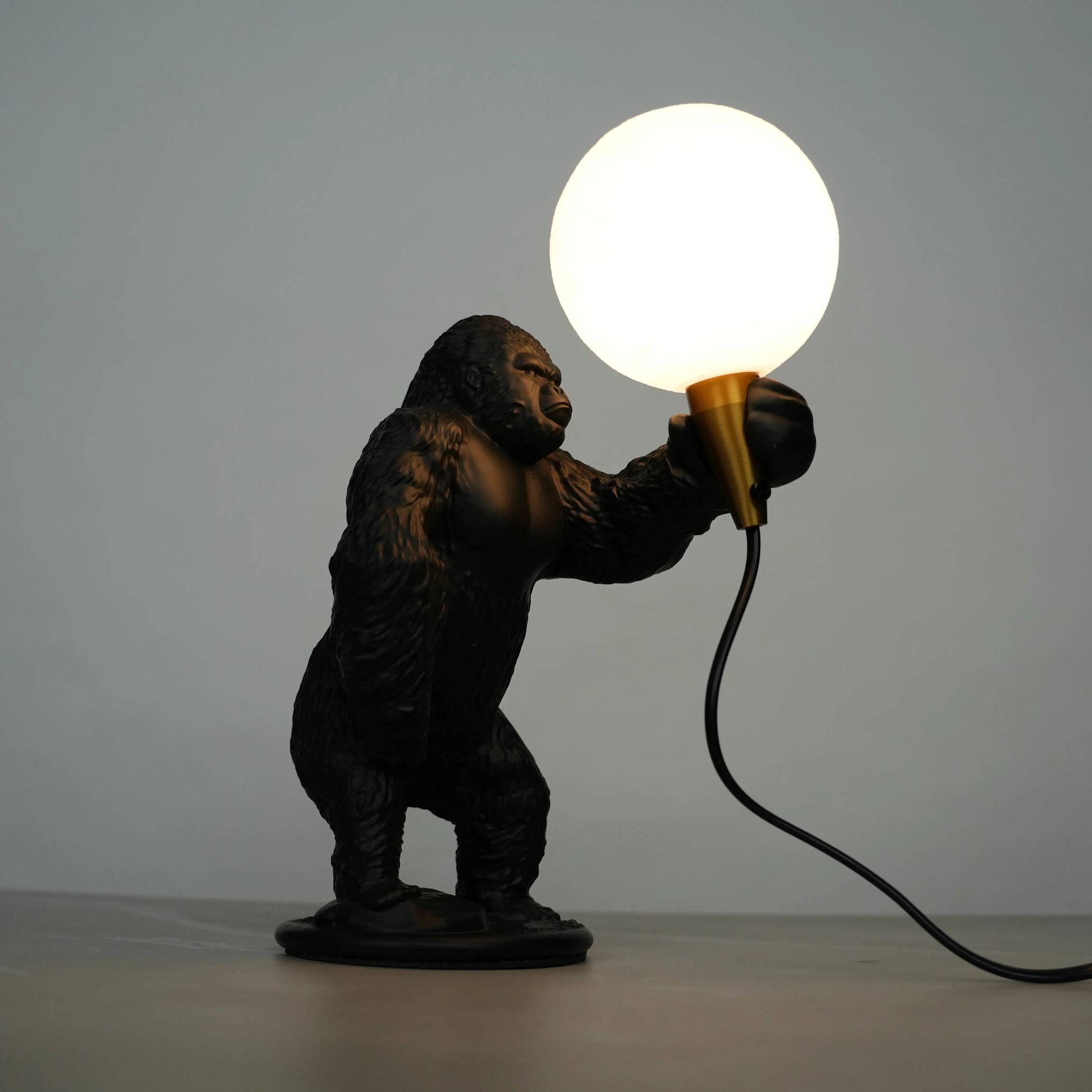 Black gorilla sculpture lamp holding a warm glowing orb with a gold lamp holder on a round black base.