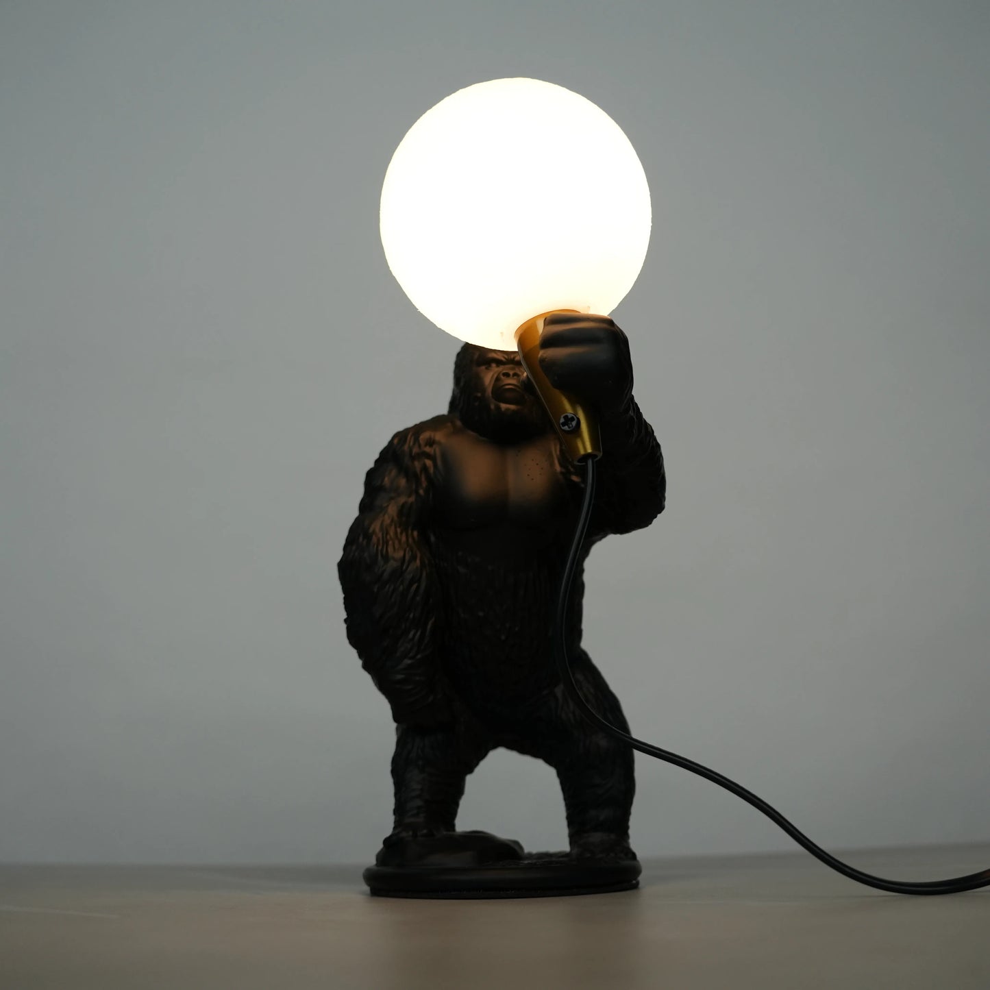 Black gorilla sculpture lamp holding a warm glowing orb with a gold lamp holder on a round black base.