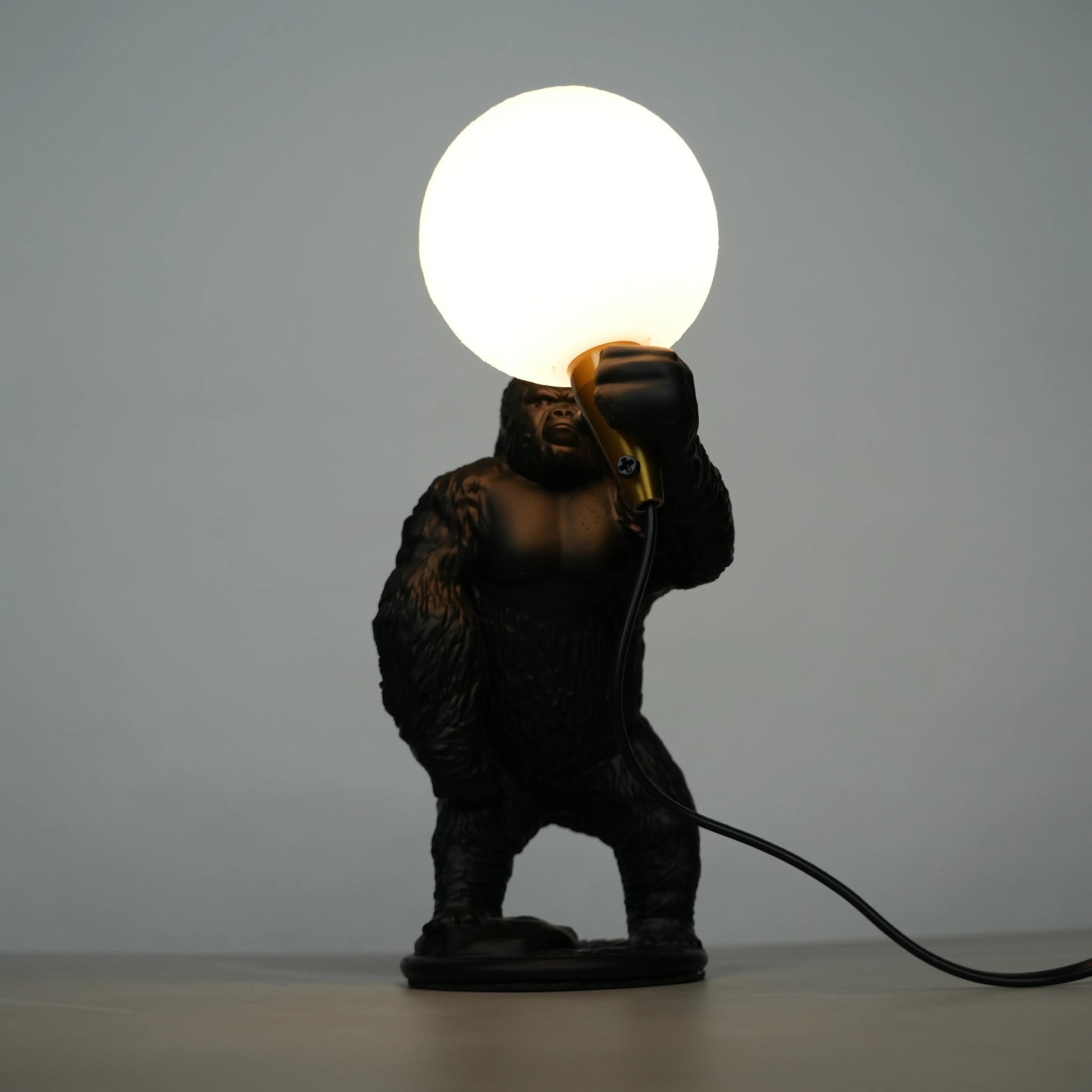 Black gorilla sculpture lamp holding a warm glowing orb with a gold lamp holder on a round black base.