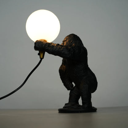 Black gorilla sculpture lamp holding a warm glowing orb with a gold lamp holder on a round black base.