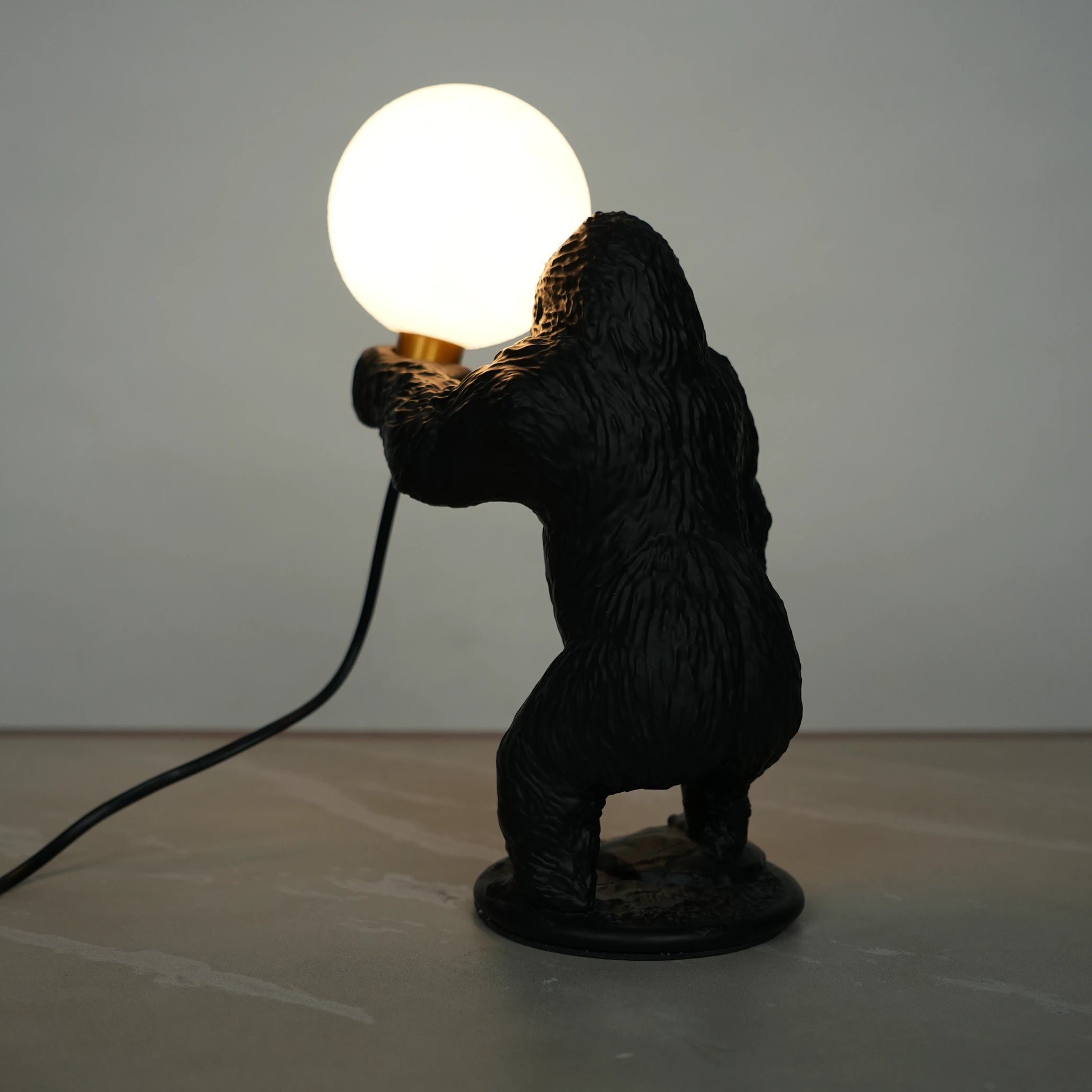 Black gorilla sculpture lamp holding a warm glowing orb with a gold lamp holder on a round black base.