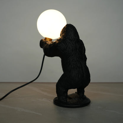 Black gorilla sculpture lamp holding a warm glowing orb with a gold lamp holder on a round black base.