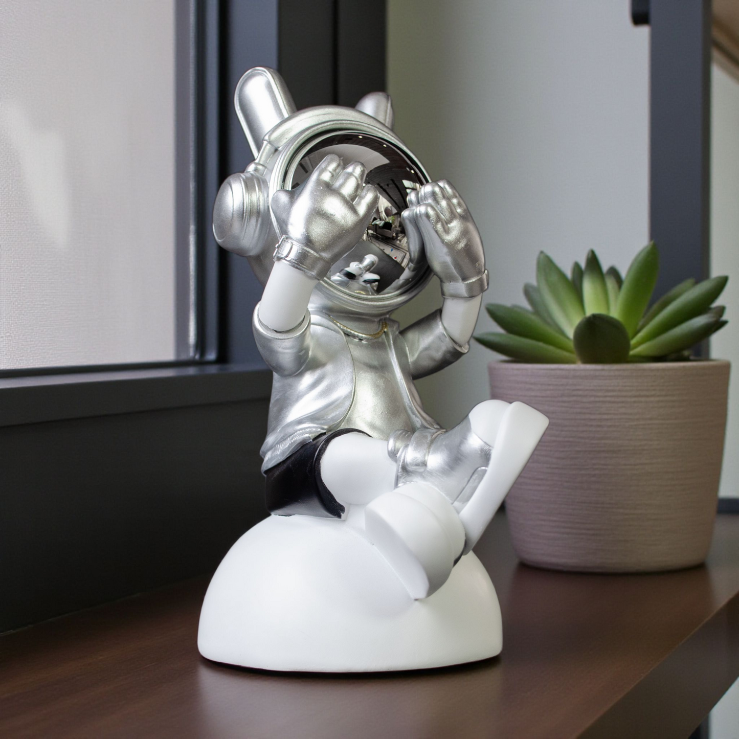 Seated astronaut figurine in silver suit covering chrome visor on white base
